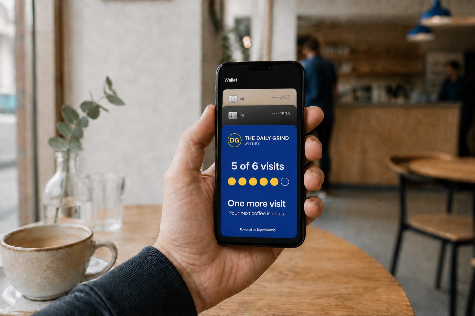 A phone held in a cafe showing a TapReward loyalty card in Apple Wallet, five of six visits collected, sitting below two bank cards